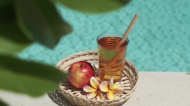 Elma suyu, bambu kamışı, kırmızı elma, tropikal çiçek frangipani, yeşil yapraklı ağaç dalı ve arka planda kaynayan mavi yüzme havuzu olan cam görüntüleri..