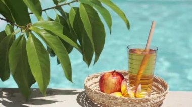 Elma suyu, bambu kamışı, kırmızı elma, tropikal çiçek frangipani, yeşil yapraklı ağaç dalı ve arka planda kaynayan mavi yüzme havuzu olan cam görüntüleri..