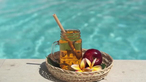 ビデオ映像のガラス製のメイソンジャーでアップルジュース 竹のわら 赤いリンゴ 熱帯の花Frangipaniと背景にバブルブルースイミングプール — ストック動画