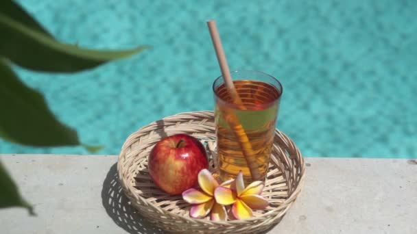 リンゴジュースとガラスのビデオ映像 竹のわら 赤いリンゴ 熱帯の花Frangipani 緑の葉を持つ木の枝と背景にバブルブルースイミングプール — ストック動画