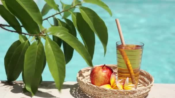 リンゴジュースとガラスのビデオ映像 竹のわら 赤いリンゴ 熱帯の花Frangipani 緑の葉を持つ木の枝と背景にバブルブルースイミングプール — ストック動画