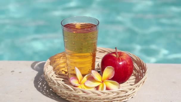 ビデオ映像のガラスとアップルジュース 竹のわら 赤いリンゴ 熱帯の花Frangipaniと背景にバブルブルースイミングプール — ストック動画