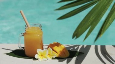 Mango suyu, bambu kamışı, taze mango, sarı frangipani çiçeği, palmiye ağacının gölgesi ve arka planda kaynayan mavi yüzme havuzu olan cam mason kavanozunun video görüntüleri..