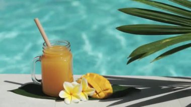 Mango suyu, bambu kamışı, taze mango, sarı frangipani çiçeği, palmiye ağacının gölgesi ve arka planda kaynayan mavi yüzme havuzu olan cam mason kavanozunun video görüntüleri..