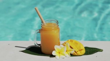 Mango suyu, bambu kamışı, taze mango, sarı frangipani çiçeği ve arka planda kaynayan mavi yüzme havuzu olan cam mason kavanozunun video görüntüleri..
