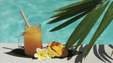 Mango suyu, bambu kamışı, taze mango, sarı frangipani çiçeği, palmiye ağacının gölgesi ve arka planda kaynayan mavi yüzme havuzu olan cam mason kavanozunun video görüntüleri..