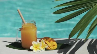 Mango suyu, bambu kamışı, taze mango, sarı frangipani çiçeği, palmiye ağacının gölgesi ve arka planda kaynayan mavi yüzme havuzu olan cam mason kavanozunun video görüntüleri..
