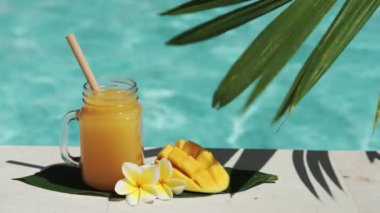 Mango suyu, bambu kamışı, taze mango, sarı frangipani çiçeği, palmiye ağacının gölgesi ve arka planda kaynayan mavi yüzme havuzu olan cam mason kavanozunun video görüntüleri..