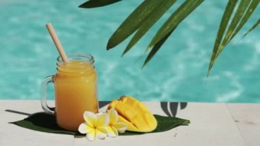Mango suyu, bambu kamışı, taze mango, sarı frangipani çiçeği, palmiye ağacının gölgesi ve arka planda kaynayan mavi yüzme havuzu olan cam mason kavanozunun video görüntüleri..