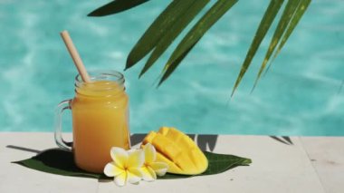 Mango suyu, bambu kamışı, taze mango, sarı frangipani çiçeği, palmiye ağacının gölgesi ve arka planda kaynayan mavi yüzme havuzu olan cam mason kavanozunun video görüntüleri..