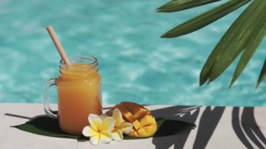 Mango suyu, bambu kamışı, taze mango, sarı frangipani çiçeği, palmiye ağacının gölgesi ve arka planda kaynayan mavi yüzme havuzu olan cam mason kavanozunun video görüntüleri..