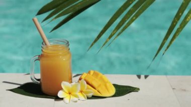 Mango suyu, bambu kamışı, taze mango, sarı frangipani çiçeği, palmiye ağacının gölgesi ve arka planda kaynayan mavi yüzme havuzu olan cam mason kavanozunun video görüntüleri..