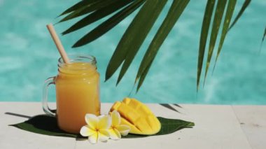 Mango suyu, bambu kamışı, taze mango, sarı frangipani çiçeği, palmiye ağacının gölgesi ve arka planda kaynayan mavi yüzme havuzu olan cam mason kavanozunun video görüntüleri..