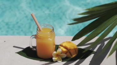Mango suyu, bambu kamışı, taze mango, sarı frangipani çiçeği, palmiye ağacının gölgesi ve arka planda kaynayan mavi yüzme havuzu olan cam mason kavanozunun video görüntüleri..