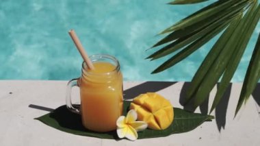 Mango suyu, bambu kamışı, taze mango, sarı frangipani çiçeği, palmiye ağacının gölgesi ve arka planda kaynayan mavi yüzme havuzu olan cam mason kavanozunun video görüntüleri..