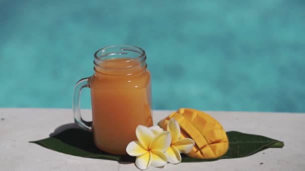 带芒果汁 一半新鲜芒果 黄色乳酪花和气泡蓝色游泳池的玻璃瓶视频镜头 — 图库视频影像