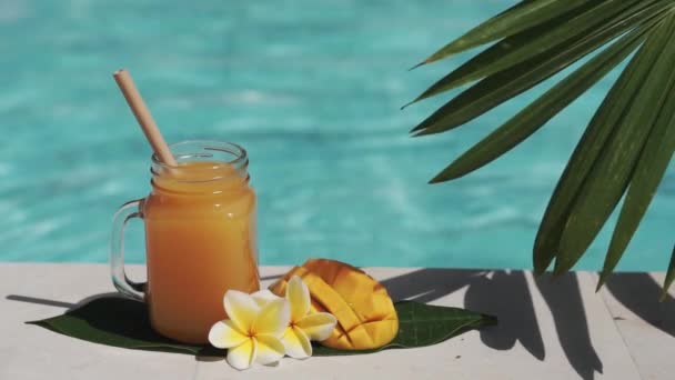 マンゴージュース 竹わら 新鮮なマンゴーの半分 黄色のフランジパニの花 ヤシの木からの日陰 背景にバブルブルースイミングプールとガラスのマソン瓶のビデオ映像 — ストック動画