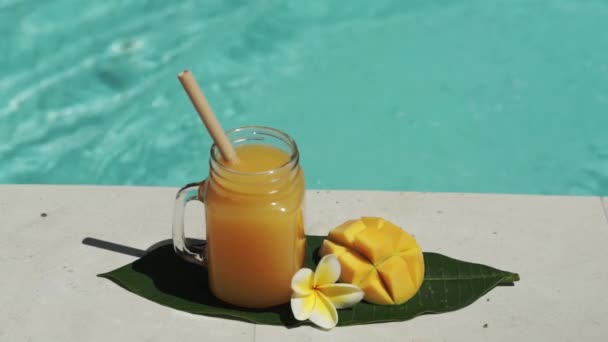 带芒果汁 一半新鲜芒果 黄色乳酪花和气泡蓝色游泳池的玻璃瓶视频镜头 — 图库视频影像