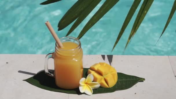 有芒果汁 一半新鲜芒果 棕榈树树荫和背景上起泡的蓝色游泳池的玻璃罐视频镜头 — 图库视频影像