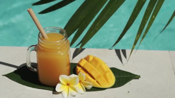 有芒果汁 一半新鲜芒果 棕榈树树荫和背景上起泡的蓝色游泳池的玻璃罐视频镜头 — 图库视频影像