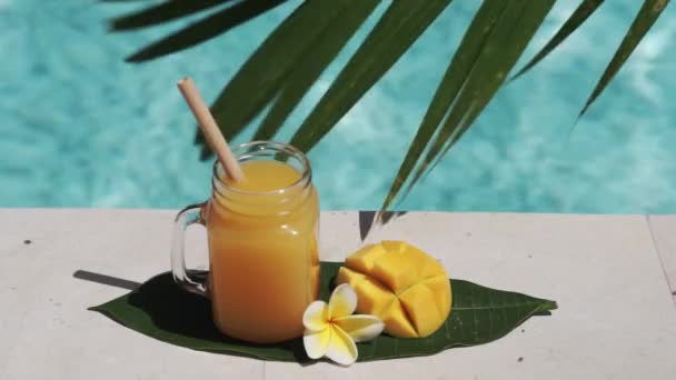 Video footage of glass mason jar with mango juice, bamboo straw, half of fresh mango, yellow frangipani flower, shade from palm tree and bubbling blue swimming pool on background.