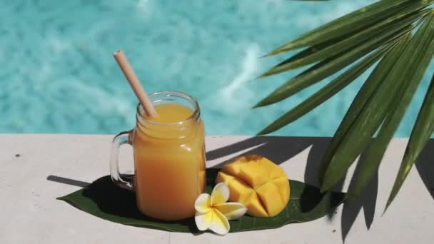 マンゴージュース 竹わら 新鮮なマンゴーの半分 黄色のフランジパニの花 ヤシの木からの日陰 背景にバブルブルースイミングプールとガラスのマソン瓶のビデオ映像 — ストック動画