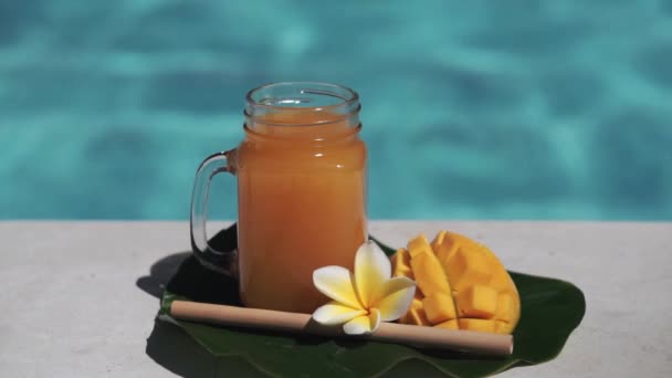 マンゴージュース 新鮮なマンゴーの半分 黄色Frangipani花と背景にバブルブルースイミングプールとガラスのマソン瓶のビデオ映像 — ストック動画
