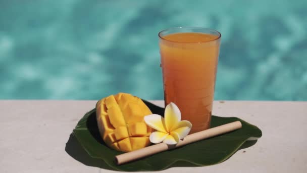 带芒果汁 一半新鲜芒果 黄色乳酪花和气泡蓝色游泳池的玻璃杯录像带 — 图库视频影像