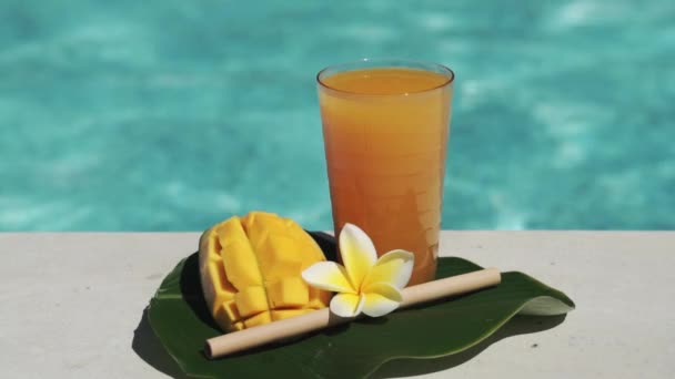 带芒果汁 一半新鲜芒果 黄色乳酪花和气泡蓝色游泳池的玻璃杯录像带 — 图库视频影像