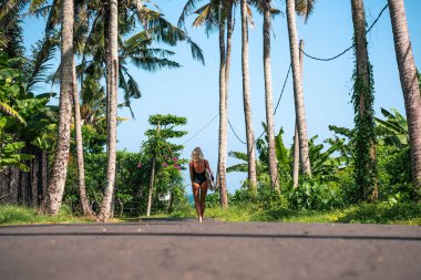 Yüzme takımlı sörfçü kız sörf tahtasıyla okyanusa doğru yürüyor, güneşli bir günde palmiye ağaçları ve Bali 'de mavi gökyüzü.