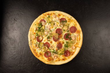 Salamlı, sosisli, salatalık turşulu, dereotlu ve peynirli taze pişmiş pizza siyah taştan bir mutfak masasında yatıyor.