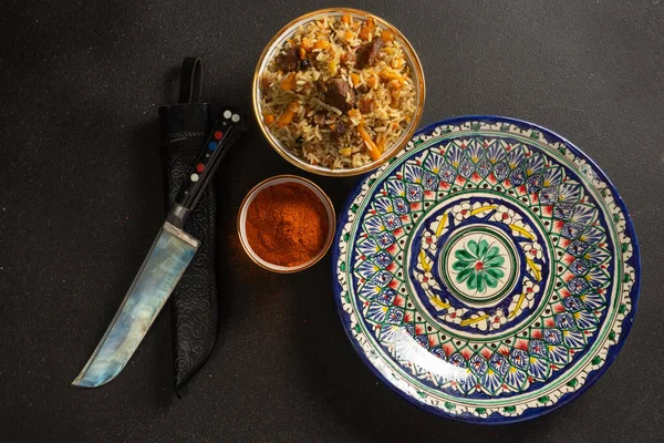 Pilaf is a traditional rice dish with lamb or beef and vegetables in ethnic Uzbek ceramic dishes. A kitchen knife with a traditional Uzbek pattern and spices.