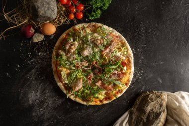 Klasik domuz pastırmalı, roka ve rendelenmiş parmesan peynirli ince hamurlu pizza. Pizza, sebze ve otlar siyah taştan mutfak masasında..