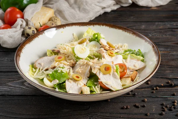 Caesar salad with chicken fillet. Lettuce, olives, cheese, cherry tomatoes, quail egg, and crunchy croutons in a ceramic plate on a wooden kitchen table. Cold appetizer. — Stock Image Caesar Salad Chicken Fillet Lettuce Olives Cheese Cherry Tomatoes Quail Royalty Free Stock Photos
