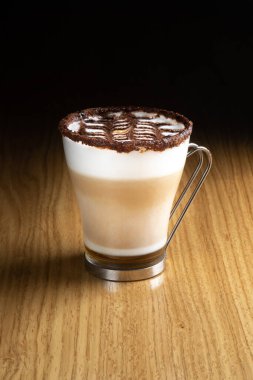 Ahşap bir masada metal saplı şeffaf bir bardakta şerbetli, sütlü ve çikolatalı Latte sanat kahvesi.