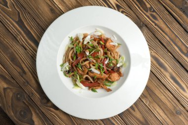 Tavuklu Çin eriştesi dolmalık biber ve teriyaki soslu soğan. Ahşap mutfak masasında beyaz seramik tabakta sıcak Asya makarnası ve kümes hayvanı eti. Üst görünüm