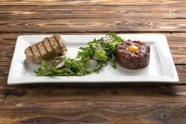 Ahşap mutfak masasında seramik tabakta salamura edilmiş sığır etli tartar, bıldırcın yumurtası, marul, turp ve ızgara ciabatta..