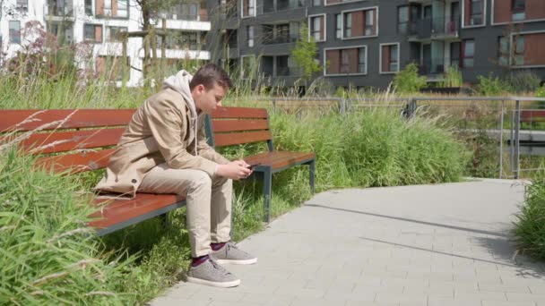 Un tipo triste está escribiendo por teléfono, sentado en un banco cerca de la casa — Vídeos de Stock