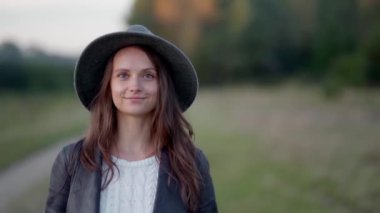 Şapkalı bir kadın parkta yürüyor, kameraya bakıyor ve hafifçe gülümsüyor.