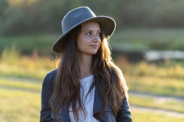 Genç, güzel, uzun saçlı, şapkalı, gülümseyen bir kadın gün batımının ışıltısında parktaki kameraya bakıyor.