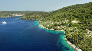 A beautiful Croatian bay with deep blue and turquoise water surrounded by a lush pine forest. A luxury yacht is anchored in the calm sea near a small coastal village in this wide aerial view.