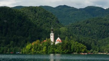 A scenic view of the iconic Pilgrimage Church of the Assumption of Mary on a small island in Lake Bled, Slovenia. This famous landmark is surrounded by calm water and dense green forest hills.