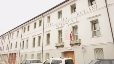 PORDENON, İTALYA - 3 Kasım 2025: yaşlılar için Anziani Umberto I Bakımevi 'nin dış cephesi. Yağmurlu bir günde İtalyan bayrağıyla beyaz bir bina ve park edilmiş arabalar..