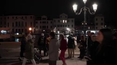 VENEZIA, İtalya - 3 Kasım 2025: Büyük Kanal boyunca mavi acil durum ışıkları hızlanan bir polis botu olarak gece gezinti alanı boyunca yürüyen bir kalabalık. Tarihi binaların arka planı.