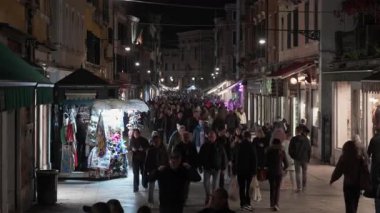 VENEZIA, İtalya - 3 Kasım 2025: Venedik 'te kalabalık dar bir cadde, açık kafelerde yürüyen ve yemek yiyen birçok insan. Kentteki tarihi binalar ve yerel dükkanlar aydınlatıldı.