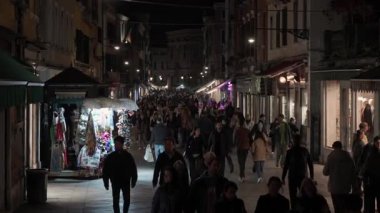VENICE, ITALY NOVEMBER 5, 2025: Meşgul alışveriş caddesi boyunca yürümekte olan yayalar, tarihi şehir merkezinde akşam turizmi sırasında ışıklı vitrinler ve hediyelik eşya kulübeleriyle.
