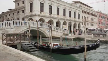 Klasik siyah gondol teknesi Venice İtalya 'da bir rıhtıma demirlemiş. Turistler bulutlu bir günde tarihi mimari ve tarihi binaların önündeki taş köprüden geçiyorlar..