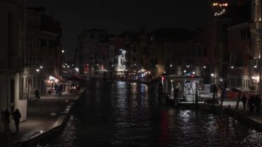 Karanlık suyu yansıtan sokak ışıklarıyla Venedik kanalının gece görüşü. İnsanlar açık bir kafe ve tekne istasyonunun önünden geçiyorlar. Tarihi İtalyan mimarisi ve şehir akşam atmosferi.