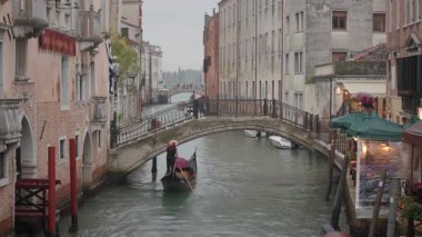 Gondolcular, yağmur sırasında taş köprü altında şemsiye tutan siyah gondolları sıralıyor. İtalya, Venedik 'te yağmurlu bir günde ıslak tarihi binalar ve dar kanallar.