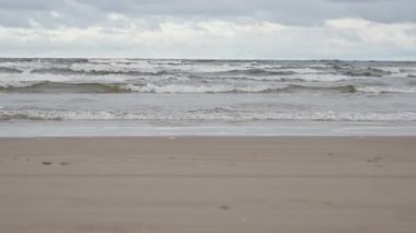 Deniz dalgaları bulutlu gri gökyüzünün altında kumlu sahil sahillerinde dalgalanır. Baltık deniz kıyısı manzarası, su hareketi ve doğa arka planıyla. Sonbahar mevsiminde deniz kenarında soğuk rüzgarlı hava.